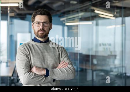 Confident bearded businessman wearing eyeglasses and a casual sweater, standing with crossed arms in a contemporary corporate office, portraying professionalism and leadership Stock Photo