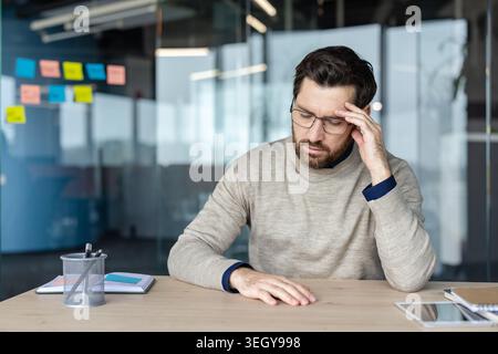 Exhausted stressed businessman sitting at desk, touching temples, suffering from headache and overwork, feeling pressure and frustration in a modern office environment Stock Photo
