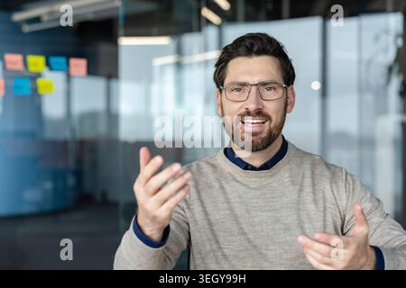 Professional man wearing glasses and a beard smiling while explaining a complex concept using hand gestures during a business meeting or online video call Stock Photo