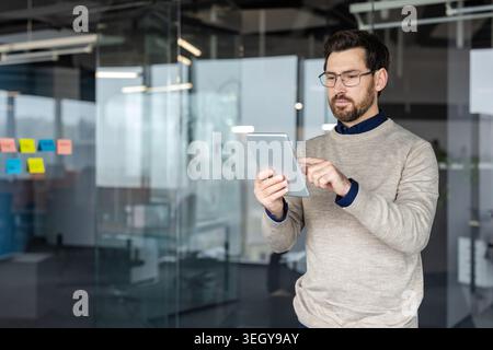 Professional man wearing glasses and a sweater standing in a contemporary office, focusing on his wireless digital tablet while interacting with the touchscreen device Stock Photo