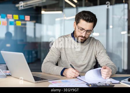 Professional man at a modern office desk reviewing documents, signing papers and analyzing finances with laptop, calculator and charts for planning and management decisions Stock Photo