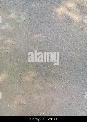 Asphalt Pavement Displaying Faint Dappled Sunlight And Subtle Shadow Patterns On Outdoor Path Surface With Fine Gravel And Textured Granules During Bright Daytime. Stock Photo