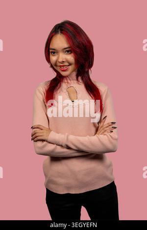 Young woman smiling while standing with her arms crossed on a pink studio background Stock Photo