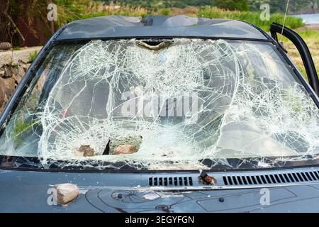 Damaged vehicle featuring a smashed front windshield and heavy rock impact on the hood. Scene depicts severe property damage from an accident or vanda Stock Photo