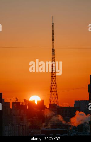 Cityscape featuring a tall communications tower piercing the bright orange sky, with the setting sun illuminating a horizon of buildings releasing plumes of smoke, portraying a modern metropolis Stock Photo