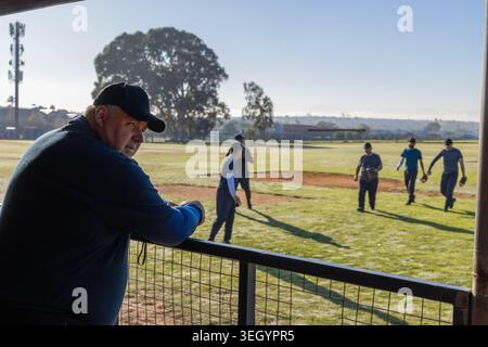 Senior man leaning on metal fence at baseball field watching male players with gloves, copy space Stock Photo
