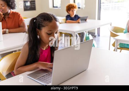 Classroom children with instructor using silver laptop and tablet in bright room, girl wearing pink Stock Photo