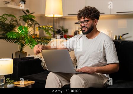 Arabian man at home opens laptop launches project types fast works remote smiles focused. Egyptian guy on sofa freelances answers email edits files chats with client writes code updates docs on time. Stock Photo