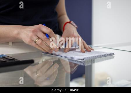 Woman Writing Notes on Glass Desk. Close-up of a woman writing in a spiral notebook on a reflective glass desk with calculator nearby. Stock Photo