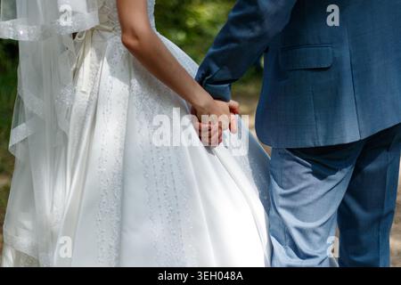 A couple walks hand in hand on a path surrounded by trees. They are dressed in formal wedding attire. Sunlight filters through the leaves above. The s Stock Photo
