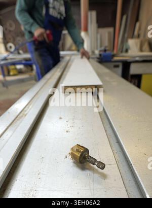Carpenter operating woodworking machine, preparing white laminated board amid drill bit and sawdust in professional workshop. 2026, 2027 Stock Photo