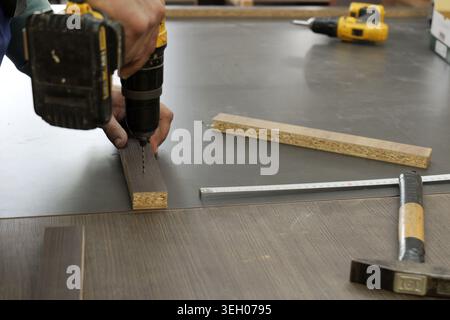 Person's hands using a cordless electric drill to create a hole in a wooden plank, working on furniture assembly or home improvement project. 2026, 20 Stock Photo