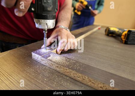 Craftsman using a cordless electric drill to create a pilot hole in a manufactured wood board, ensuring precise furniture assembly in a workshop. 2026 Stock Photo