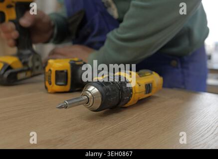 Worker placing cordless drill with screwdriver bit on light wooden table, blurred drill held in background for renovation. 2026, 2027 Stock Photo