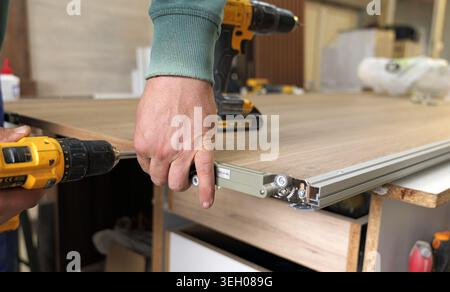 Carpenter's hands using a yellow cordless drill to securely fasten a mechanism onto a wooden furniture panel, illustrating craftsmanship and home impr Stock Photo