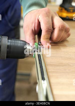 Carpenter's hand using an electric drill, screwing a drawer slide into a wooden furniture panel, illustrating craftsmanship, diy assembly, and woodwor Stock Photo