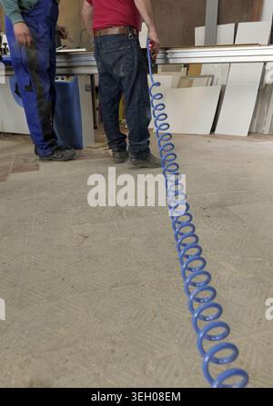 Two men in blue overalls stand in a busy woodworking shop handling an air compressor hose for cleaning and operating machinery amid dusty floors, tool Stock Photo