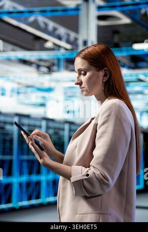 Data center female engineer using tablet app to evaluate equipment diagnostic reports. Woman in server hub scripting automated workflows using touchscreen device to oversee mainframes Stock Photo