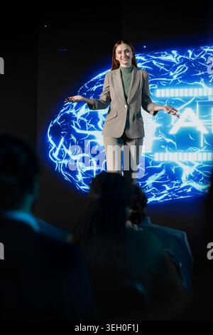 Female CEO presenter gives AI concept speech featuring AI-assistant and automation, engages investors and team during business forum in auditorium. Artificial intelligence panel discussion. Stock Photo