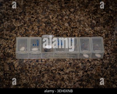 A clear plastic weekly pill box with one white tablet inside an open compartment on a dark granite counter top Stock Photo