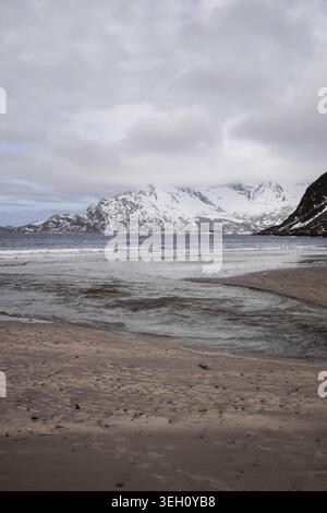 View of a sandy beach with a small stream flowing into the sea under snow-capped mountains and a cloudy sky in Kvaloya, Troms, Norway. Stock Photo