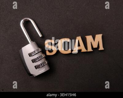 Wooden letters spelling SCAM lie next to an open silver combination padlock on a textured black surface. Stock Photo