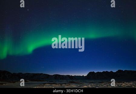 Aerial view of the Aurora Borealis glowing green over snowy mountain peaks and a frozen landscape under a starry night sky in Tasiilaq, Sermersooq Mun Stock Photo