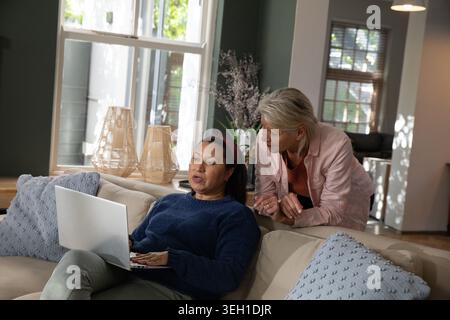 Mature female friends sitting on light-colored sofa at home, using laptop by window, wicker lamps Stock Photo