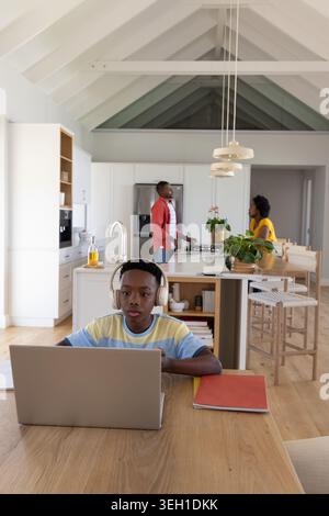 African American family watching schoolboy using laptop with headphones at kitchen table Stock Photo