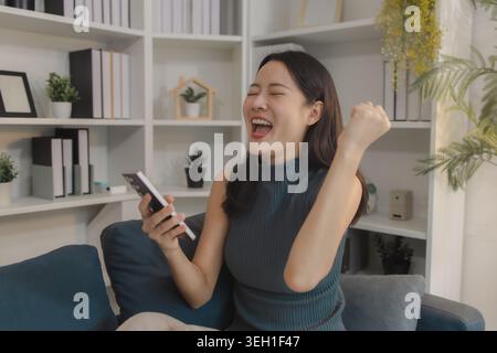 Young Asian woman sitting on sofa celebrating while using smartphone, expressing excitement and success, representing positive online experience, digi Stock Photo