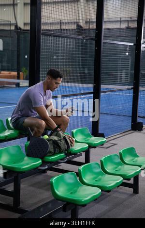 Leaning man in purple tee, shorts, scrolling phone on green seats beside padel court with paddle Stock Photo