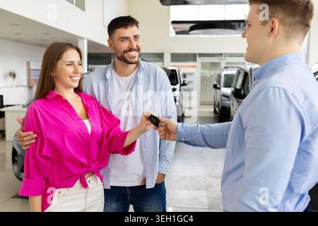 Happy young couple receiving vehicle keys from professional salesman at dealership showroom Stock Photo