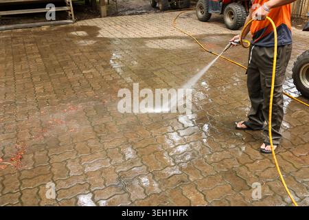 Focused worker operating high pressure washer equipment, cleaning dirty paving stone on residential driveway. Powerful water spray from yellow hose re Stock Photo