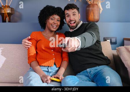 Happy multiracial couple watching television and using remote control on sofa Stock Photo