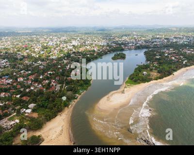 Aerial view of the Nilwala River mouth meeting the ocean with sandy beaches and urban rooftops surrounded by tropical greenery in Matara, Southern Pro Stock Photo
