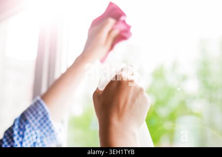 Close-up of hands holding a spray bottle and wiping a bright window with a pink cloth. Sunlit spring house cleaning concept with green bokeh backgroun Stock Photo