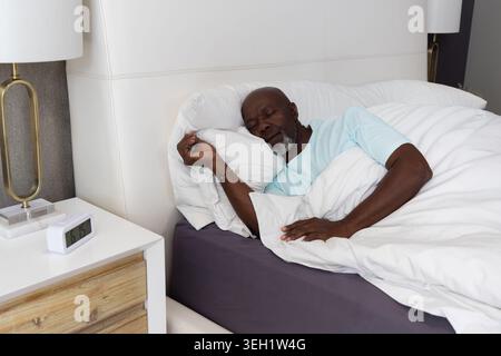 Senior African American man sleeping on right side in bedroom with pillow, duvet, lamp, alarm Stock Photo