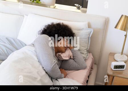 Man sleeping on right side on bed wearing light gray top with pillows, duvet, alarm clock Stock Photo