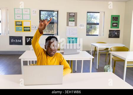 Male youth student sitting at table in class wearing yellow shirt raising hand using silver laptop Stock Photo