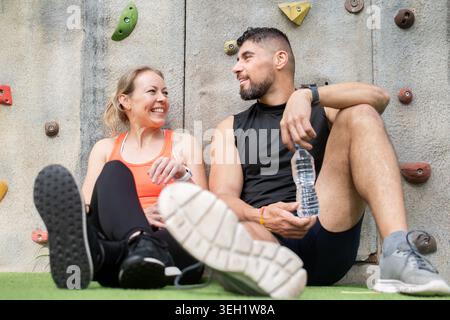 Happy athletic man and woman sitting on the ground and relaxing after exercise beside an outdoor climbing wall. Fitness, hydration, friendship and hea Stock Photo