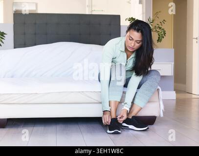 Mid-adult Asian woman sitting on bed in bedroom tying black shoes with fitness watch, copy space Stock Photo