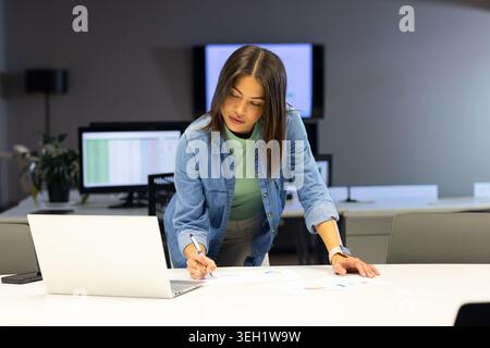 Indian woman leaning over office table, denim shirt, examining charts and using silver laptop Stock Photo