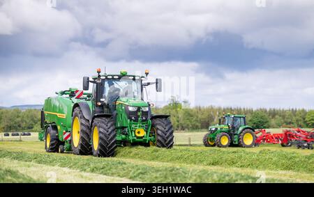 a pair of John Deere tractors, one with a rowing up machine and one with a baler, in a silage field on the edge of the Scottish Borders. Stock Photo