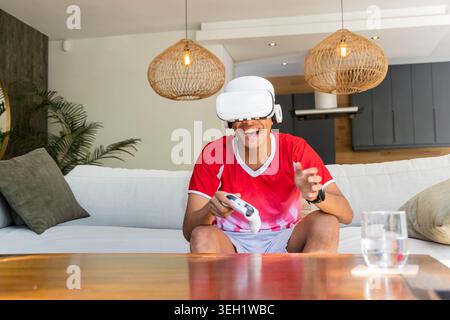 Man wearing white VR headset holding controller, laughing and leaning on sofa at home, red shirt Stock Photo
