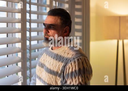 Middle-aged man standing at window at home, wearing cable-knit sweater, shutters casting shadows Stock Photo