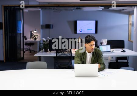 Man in green jacket working on silver laptop, reviewing printed charts at open-plan office Stock Photo