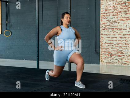 Asian woman in her 20s performing lunge in gym wearing light blue activewear near gymnastic rings Stock Photo