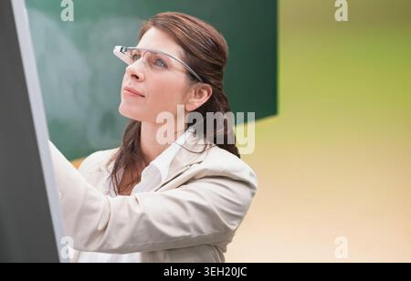 Mature woman standing near board at left, pointing at chalkboard, wearing blazer and smart glasses Stock Photo