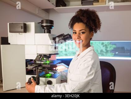 African American female scientist in lab coat using microscope with green slide at lab bench Stock Photo