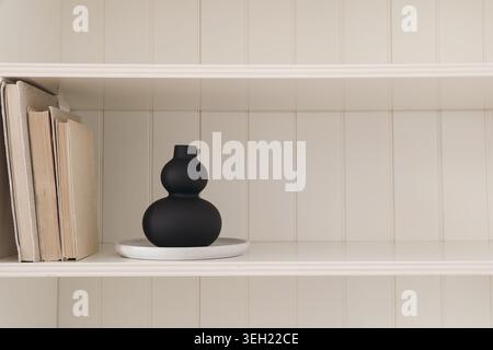 Minimalist Home Decor with Modern Organic shaped Black Ceramic Vase and Old Books on White Wooden Shelving. Scandinavian Interior Design Background Stock Photo
